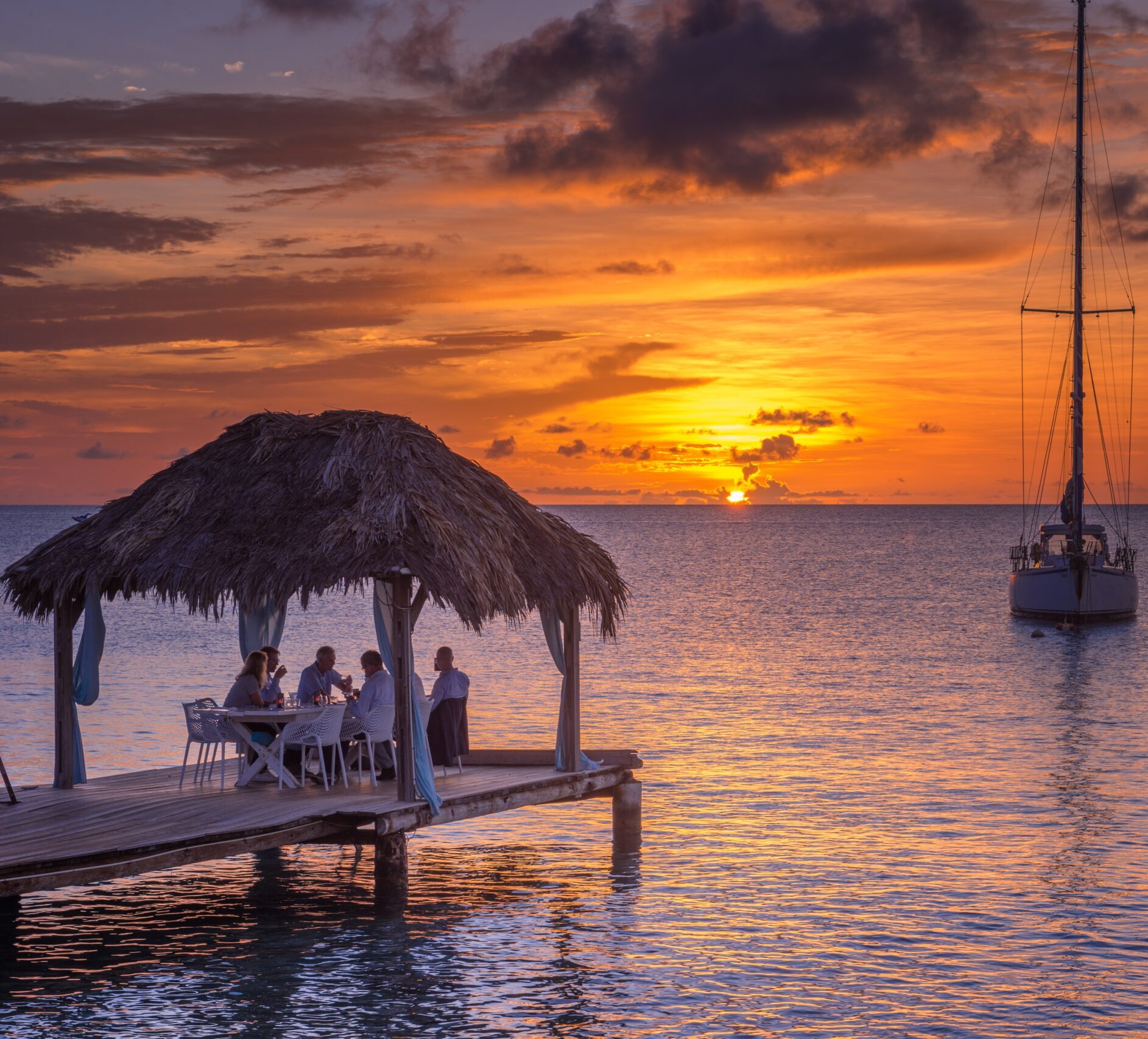 Sunset over a beach restaurant in Bonaire, tables and chairs, and the ocean in the background under a colorful orange and pink sky.